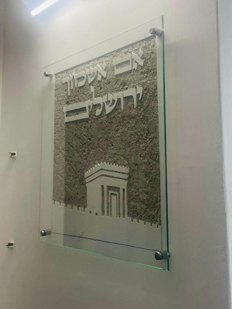 תצוגת גלריה במבט אלכסוני: אריח סיתות מרובע באבן אפורה מאחורי לוחית זכוכית שקופה על קיר לבן — נראים הכיתוב 'אם אשכחך / ירושלים' ובית המקדש מעל חומות ירושלים, וניצוץ אור משתקף משולי הזכוכית.