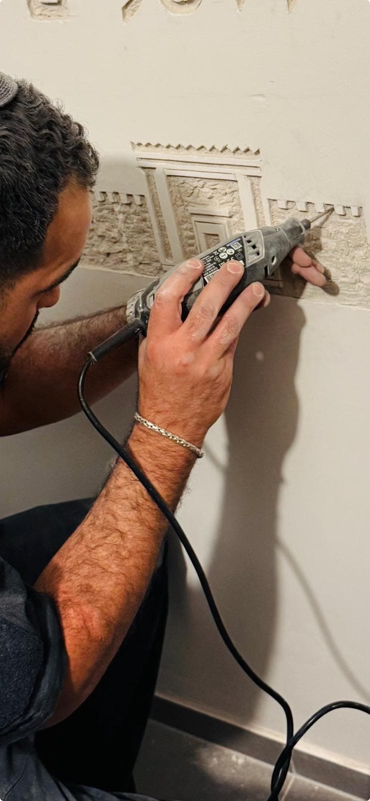 צילום פעולה: גיא, חובש כיפה, מסתת בכלי סיבובי קיר באמצע עבודה — בקיר מתגלה דמותו של בית המקדש מעל חומה משוננת של ירושלים, מסותתת מתוך הטיח כדי לחשוף את הבטון שמתחתיו.